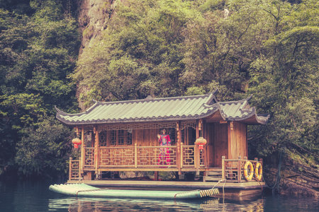 Zhangjiajie, China -  August 2019 : Chinese woman dressed in traditional ethnic minority costume standing on the porch of an old wooden floating house cabin on Baofeng Lake and waving to tourists passing by in a raft boat, Zhangjiajie National Forest Parkのeditorial素材