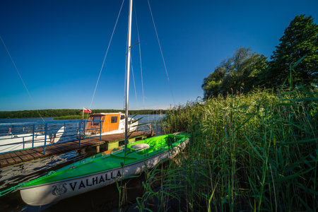 Jezioro Ostrowieckie, Poland - July 2020 : A small pier and private boats on the shore of a lake surrounded by reed and trees under the blue skyのeditorial素材