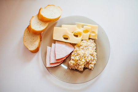 Fresh bread, meat slices, Maasdam cheese, wheat porridge on a transparent glass plate on a white tableの写真素材