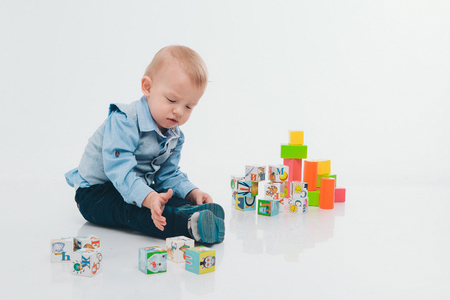 blond boy in blue suit plays with colorful blocks on white background. A small child builds a tower of cubes on the floorの写真素材