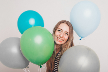 blonde girl smiling and looking at camera between multicolored balls on white background. Studio portrait of attractive girl waiting for birthday party. Face and balloonsの写真素材