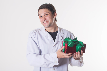 a man in a white robe holds a gift box. smiling doctor with a red box with a green bow. Handsome man looking at camera on white backgroundの写真素材
