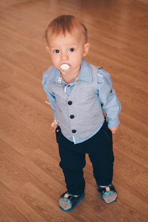 a little blond boy in a blue-and-white cosume sucks a nipple and looks into the camera standing on the floorの写真素材