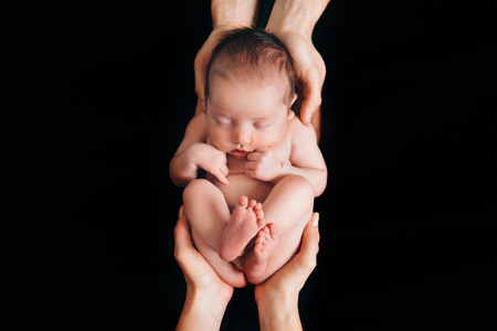 newborn baby lying on the hands of parents on a black background. Imitation of a baby in the womb. beautiful little girl sleeping lying on her backの写真素材