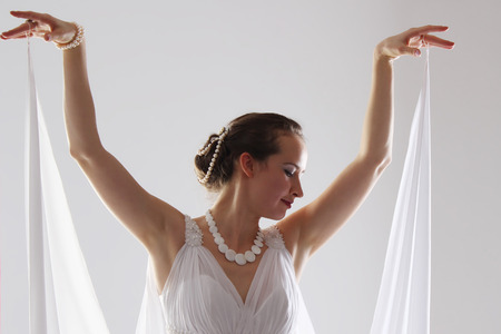 girl in white and with raised hands, attached to the floor dresses. the bride in a dress in Greek style. brunette in pearl jewelry lowered her head. The woman looks like a bird or a batの写真素材