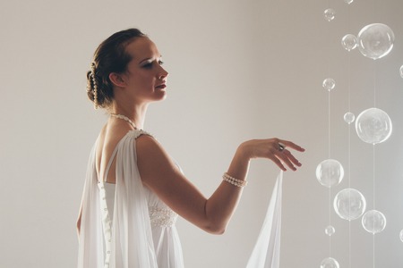 girl in white and with raised hands, attached to the floor dresses. the bride in a dress in Greek style. brunette in pearl jewelry lowered her head. The woman looks like a bird or a batの写真素材