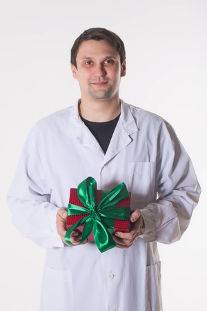 a man in a white robe holds a gift box. smiling doctor with a red box with a green bow. Handsome man looking at camera on white backgroundの写真素材