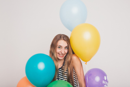 blonde girl smiling and looking at camera between multicolored balls on white background. Studio portrait of attractive girl waiting for birthday party. Face and balloonsの写真素材