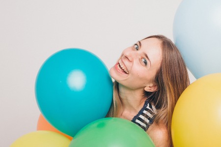 blonde girl smiling and looking at camera between multicolored balls on white background. Studio portrait of attractive girl waiting for birthday party. Face and balloonsの写真素材