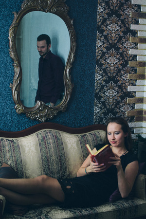 Girl in black dress reading a book on the couch, the mirror reflects the manの写真素材