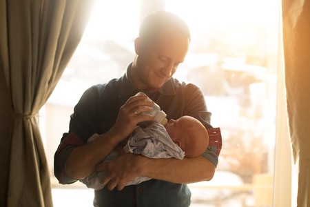 happy young father feeding newborn baby with milk bottle on couch at homeの写真素材