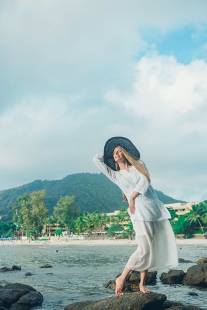 A woman in a white dress and a black hat standing on the rocks by the seaの写真素材