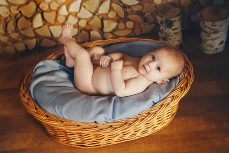 Naked baby lies in a wicker basket with his legs raised up against the background of a row of logsの写真素材