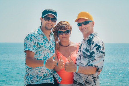 Three people in glasses stand together against the sea. Two men and a woman show a sign of pleasure with their thumbs. Family on vacationの写真素材