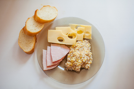 Fresh bread, meat slices, Maasdam cheese, wheat porridge on a transparent glass plate on a white tableの写真素材