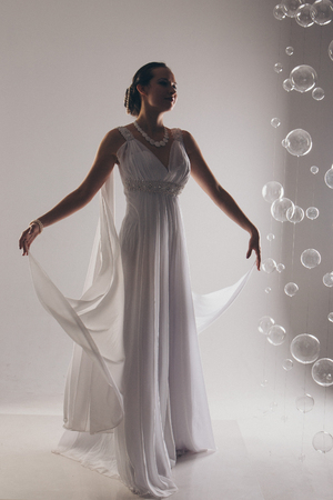 girl in white and with raised hands, attached to the floor dresses. the bride in a dress in Greek style. brunette in pearl jewelry lowered her head. The woman looks like a bird or a batの写真素材