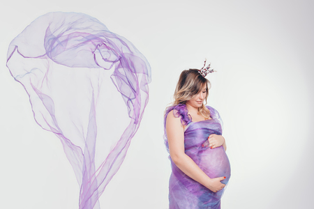 Pregnant woman in long dress with crown and flowers on white backgroundの写真素材