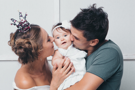 Parents hold the child in their arms between each other and kiss the baby. Man, woman in crown and daughter on white backgroundの写真素材