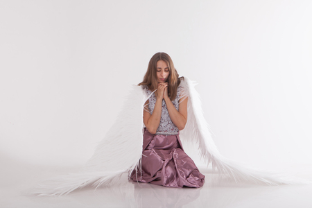 a beautiful girl with white wings is kneeling with her hands folded for prayer. Guardian Angel in pink dress on white backgroundの写真素材