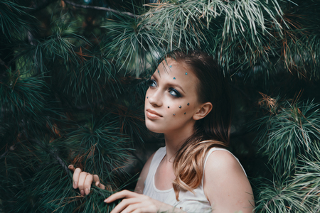 beautiful girl model with bright makeup among the branches of spruce. Portrait of a woman with rhinestones on her face close-upの写真素材