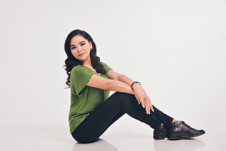 beautiful brunette posing on white background. A woman in a green shirt and black pants sitting on the floor. Model advertises clothesの写真素材