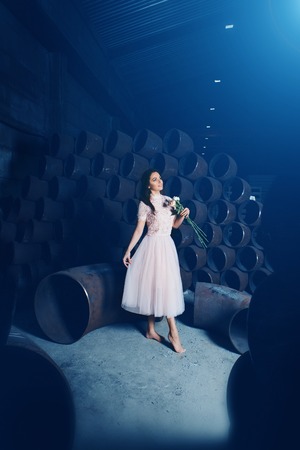 beautiful woman in light pink dress dancing barefoot with flowers on the background of large iron pipesの写真素材