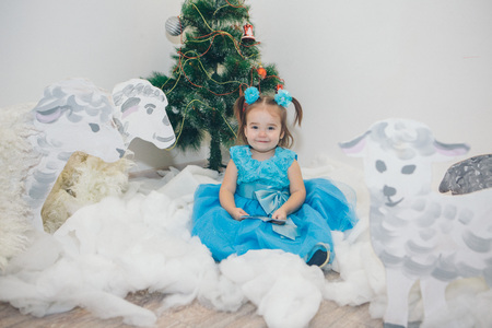 a little girl in a blue dress celebrates the new year: a child among the snow of cotton wool, painted sheep, Christmas tree and starsの写真素材