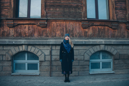 portrait of a beautiful blonde in a hat and scarf on the background of the city landscape in the cold season. girl walking down the streetの写真素材
