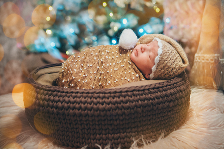 a newborn baby sleeps in a basket in hat with a POM-POM, like a gnome on the background of a Christmas tree and giftsの写真素材