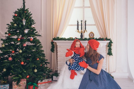 a woman with a child in a blue dress with polka dots in a red hat and mittens on the background of Christmas interior: fireplace, Christmas tree, gifts, watchesの写真素材