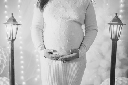 A pregnant woman in a white dress stroking her stomach near the lampposts and Christmas tree on the background of bright lightsの写真素材