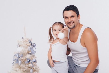 a man holds a little girl in his arms helping her dress up the Christmas tree with white balls on a white backgroundの写真素材