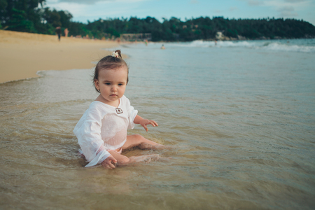 little girl in white clothes playing with sand on the beach among the wavesの写真素材