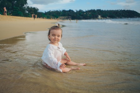 little girl in white clothes playing with sand on the beach among the wavesの写真素材