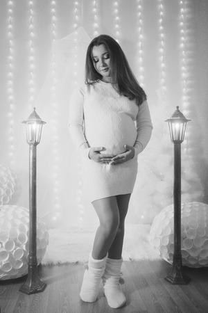 A pregnant woman in a white dress stroking her stomach near the lampposts and Christmas tree on the background of bright lightsの写真素材