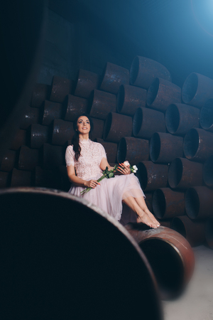 March 8: beautiful girl in a light dress barefoot with bouquet of flowers among metal pipesの写真素材