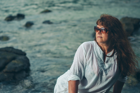 a woman in sun glasess on a rock looking into the distance against the seaの写真素材