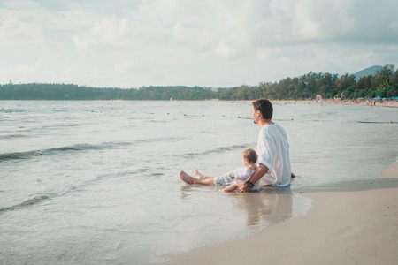 Dad in sunglasses holds the baby in his arms on the beach background. The concept of educating the father of young children, Happy childhood, a friendly family.の写真素材