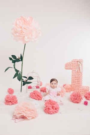 Birthday celebration: girl sitting on the floor among the numbers 1, artificial flowers and bird cageの写真素材