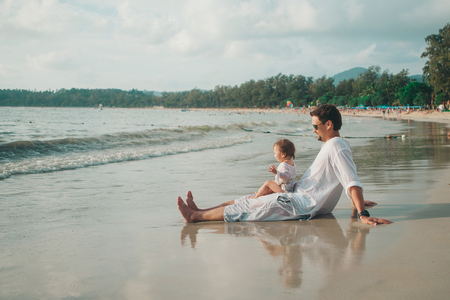 Dad in sunglasses holds the baby in his arms on the beach background. The concept of educating the father of young children, Happy childhood, a friendly family.の写真素材