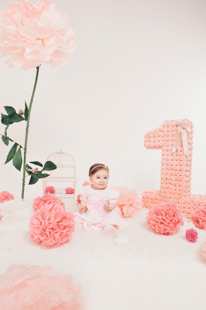 Birthday celebration: girl sitting on the floor among the numbers 1, artificial flowers and bird cageの写真素材