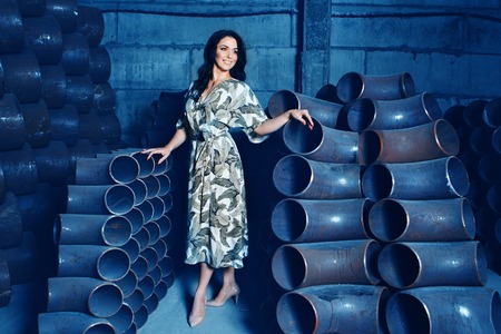 a beautiful brunette with a bright red manicure in a dress and shoes poses among the metal pipes. A woman in beautiful clothes standing in a metal warehouseの写真素材