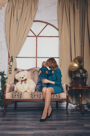 the child in the arms of his mother on the couch. A woman with a child in her arms among the decor elements: a gramophone, a big Teddy bear, curtains, glass ballsの写真素材