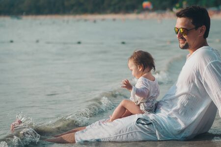 Dad in sunglasses holds the baby in his arms on the beach background. The concept of educating the father of young children, Happy childhood, a friendly family.の写真素材