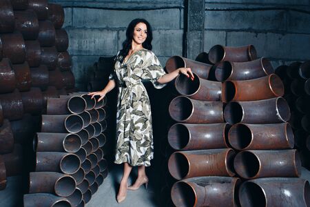 a beautiful brunette with a bright red manicure in a dress and shoes poses among the metal pipes. A woman in beautiful clothes standing in a metal warehouseの写真素材