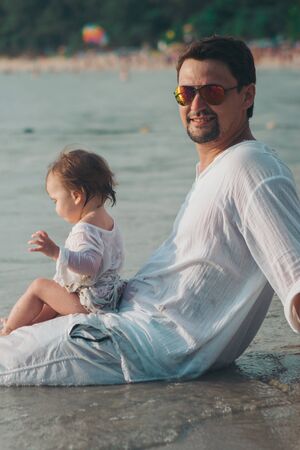 Dad in sunglasses holds the baby in his arms on the beach background. The concept of educating the father of young children, Happy childhood, a friendly family.の写真素材