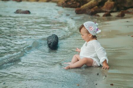 a small child sits on a sandy beach and wets his feet in the water. Girl playing in the sea on the shoreの写真素材