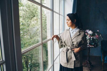 beautiful Asian woman with a hairstyle in Japanese style and a kimono at the window on the background of the home interiorの写真素材