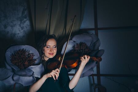 the concept of music education, the fashion Industry-a beautiful girl playing the violin among the big artificial flowers at dusk. Beauty in a dress among creative sceneryの写真素材