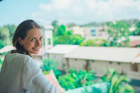 the concept of tourism, leisure is a beautiful girl with long hair, basking in the white dress on the balcony of the hotel with views of tropical plants and a small buildingの写真素材